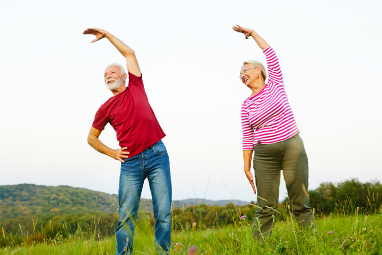 Ein älteres Paar führt Dehnübungen im Freien auf einer Wiese durch, umgeben von Landschaft und Hügeln.