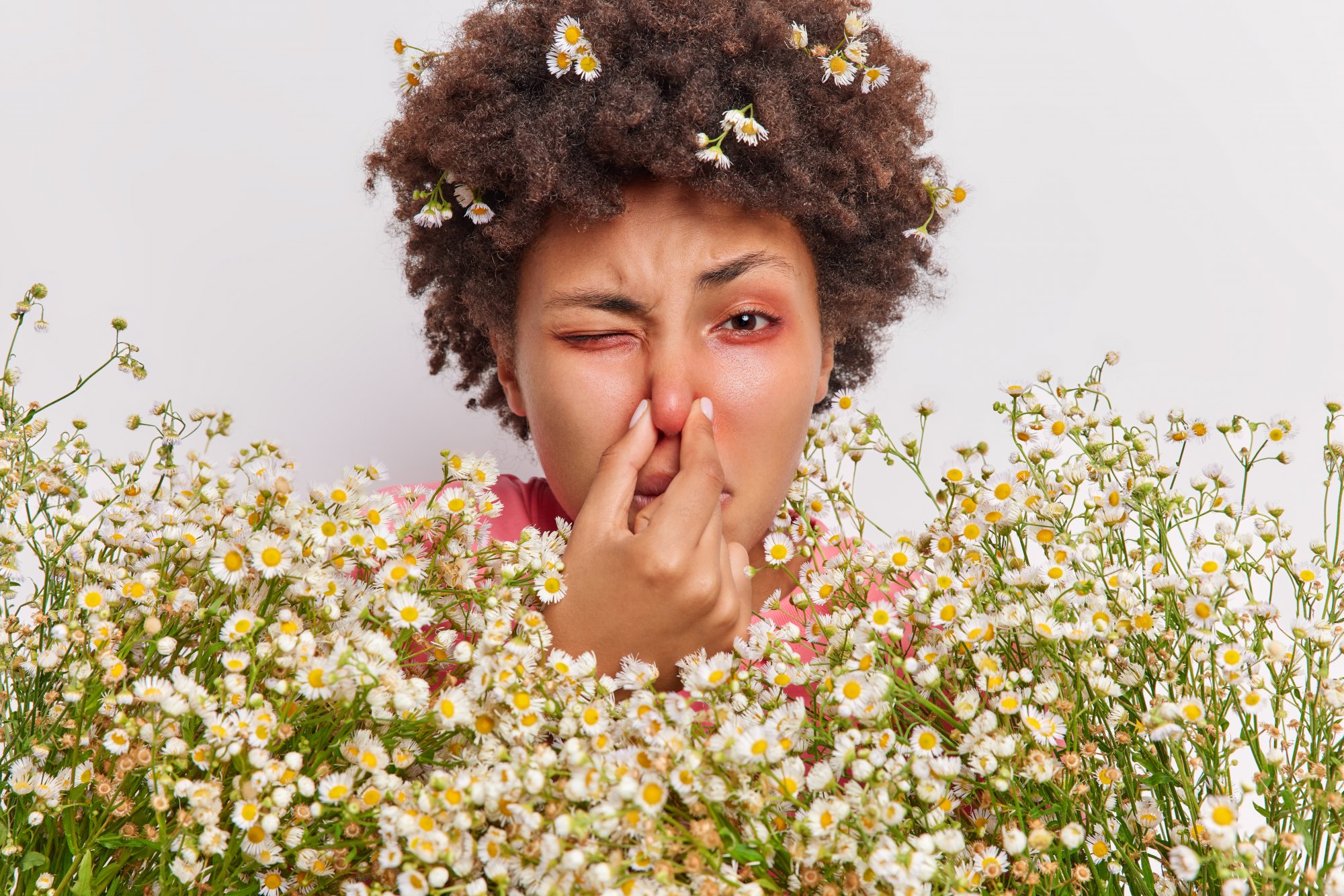 Eine Person mit lockigem Haar, die eine Blüte im Gesicht hält und aufgrund des Geruchs eine Grimasse schneidet, umgeben von Blumen.