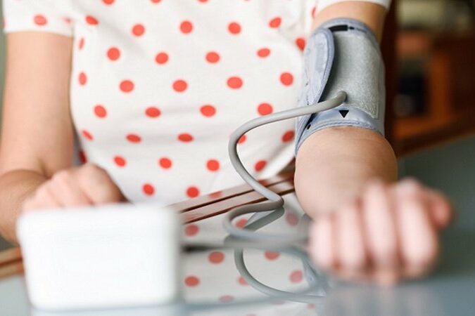 Eine Person mit einem Blutdruckmessgerät am Arm, das auf einem Tisch liegt. Im Hintergrund ein unscharfer Raum.