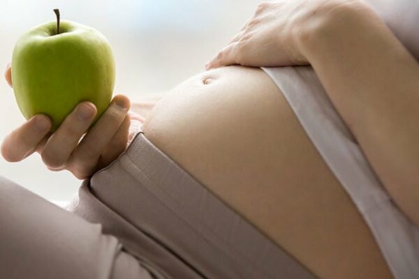 Eine schwangere Person hält einen grünen Apfel und berührt sanft ihren Bauch.