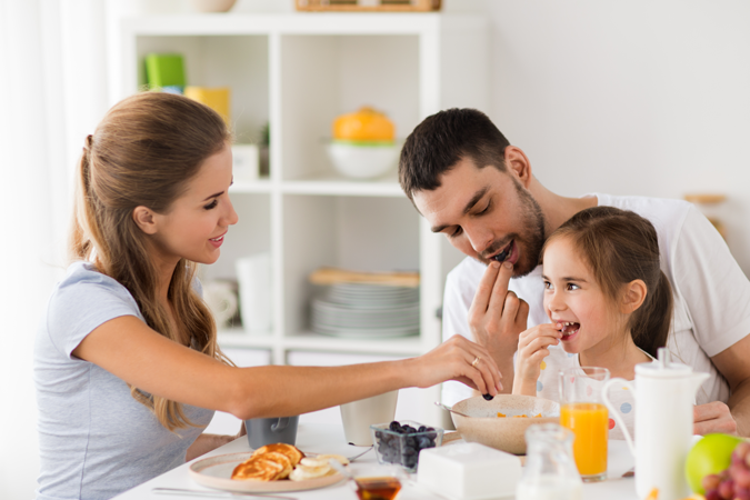 Familie beim gemeinsamen Frühstück an einem Tisch mit Obst, Pfannkuchen und Getränken.