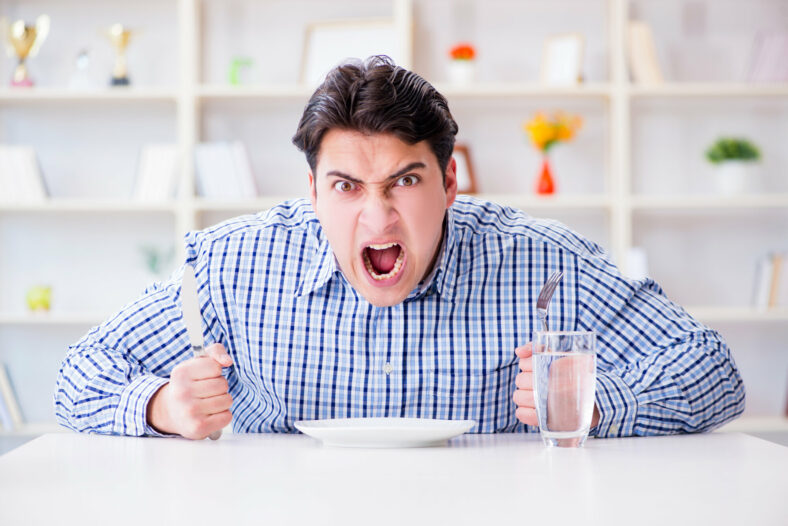 Ein wütender Mann hält ein Messer und eine Gabel über einem leeren Teller, während er an einem Tisch sitzt.