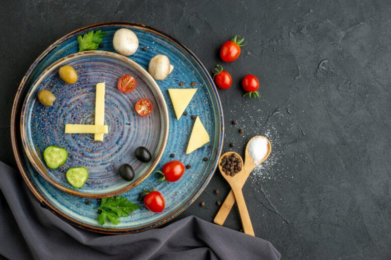 Ein kreativ angerichteter Teller mit Gemüse, Käse und Gewürzen in Form einer Uhr auf blauem Geschirr.