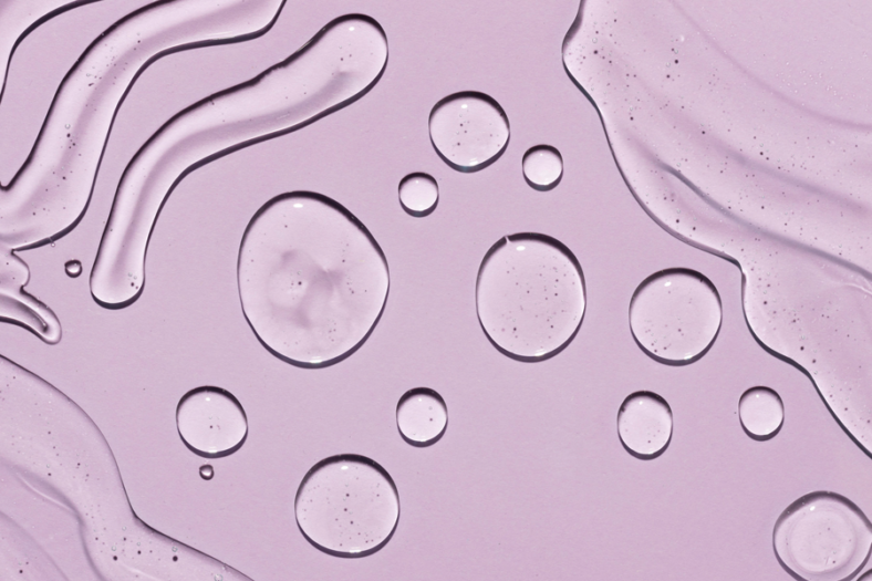 Hyaluron-web.png Transparente Wassertropfen auf einem helllila Hintergrund, mit fließenden Linien und Blasen.