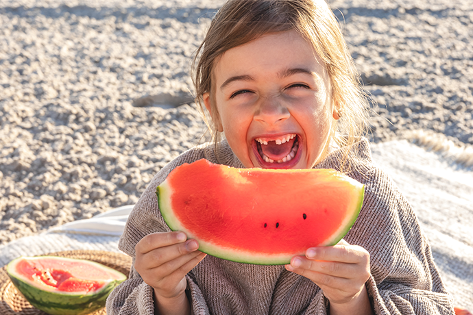 Ein lachendes Kind hält ein großes Stück Wassermelone in der Hand, umgeben von einer sommerlichen Picknick-Atmosphäre.