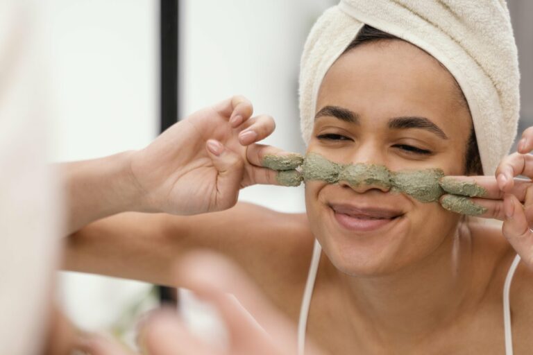 Frau mit Handtuch um den Kopf zeigt zufrieden eine Gesichtsmaske aus grünem Material im Badezimmer.