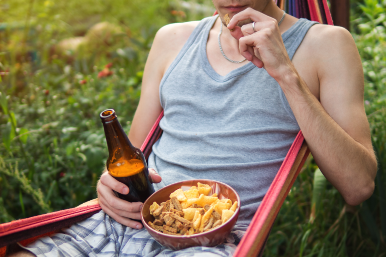 Eine Person in der Hängematte mit einer Flasche Bier und einer Schüssel Snacks, umgeben von grünen Pflanzen.