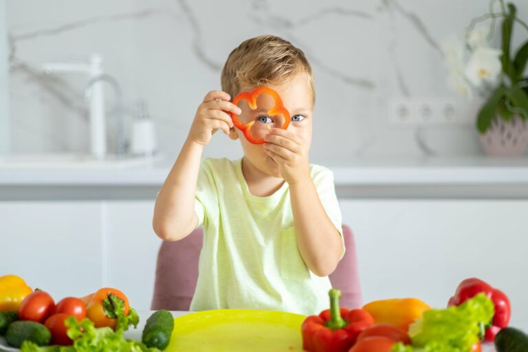 Ein Junge hält eine rote Paprika in Form eines Blumenmotivs und schaut in die Kamera, umgeben von frischem Gemüse.