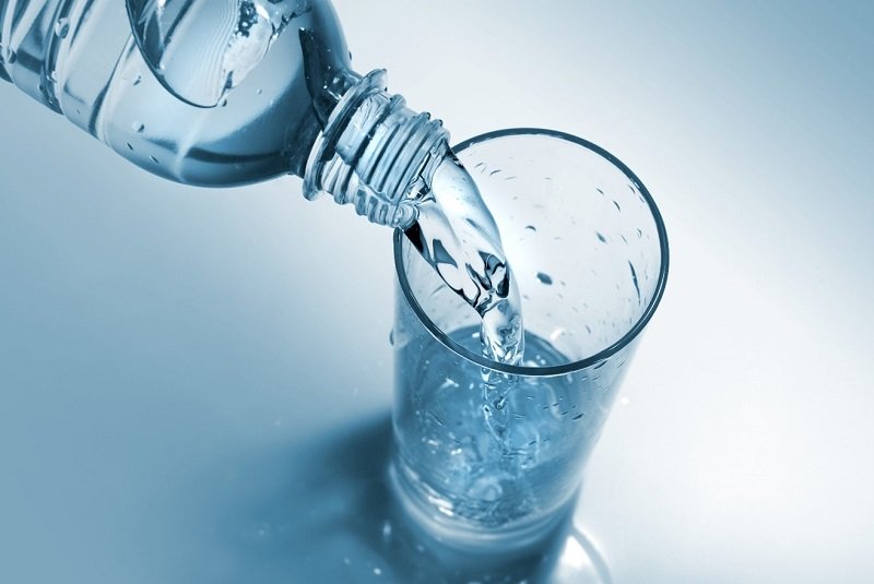 Eine Wasserflasche schüttet frisches Wasser in ein Glas, das auf einer hellen, blauen Oberfläche steht.
