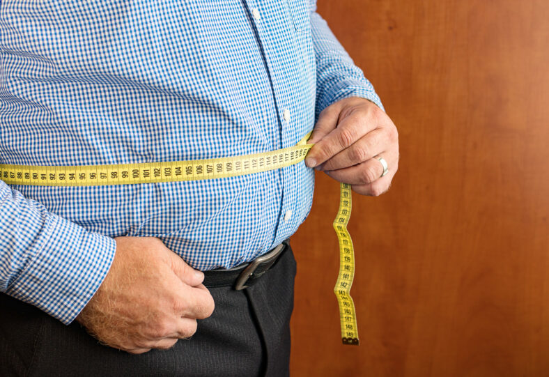 Mann misst seinen Bauchumfang mit einem gelben Maßband, trägt ein kariertes Hemd und dunkle Hose.