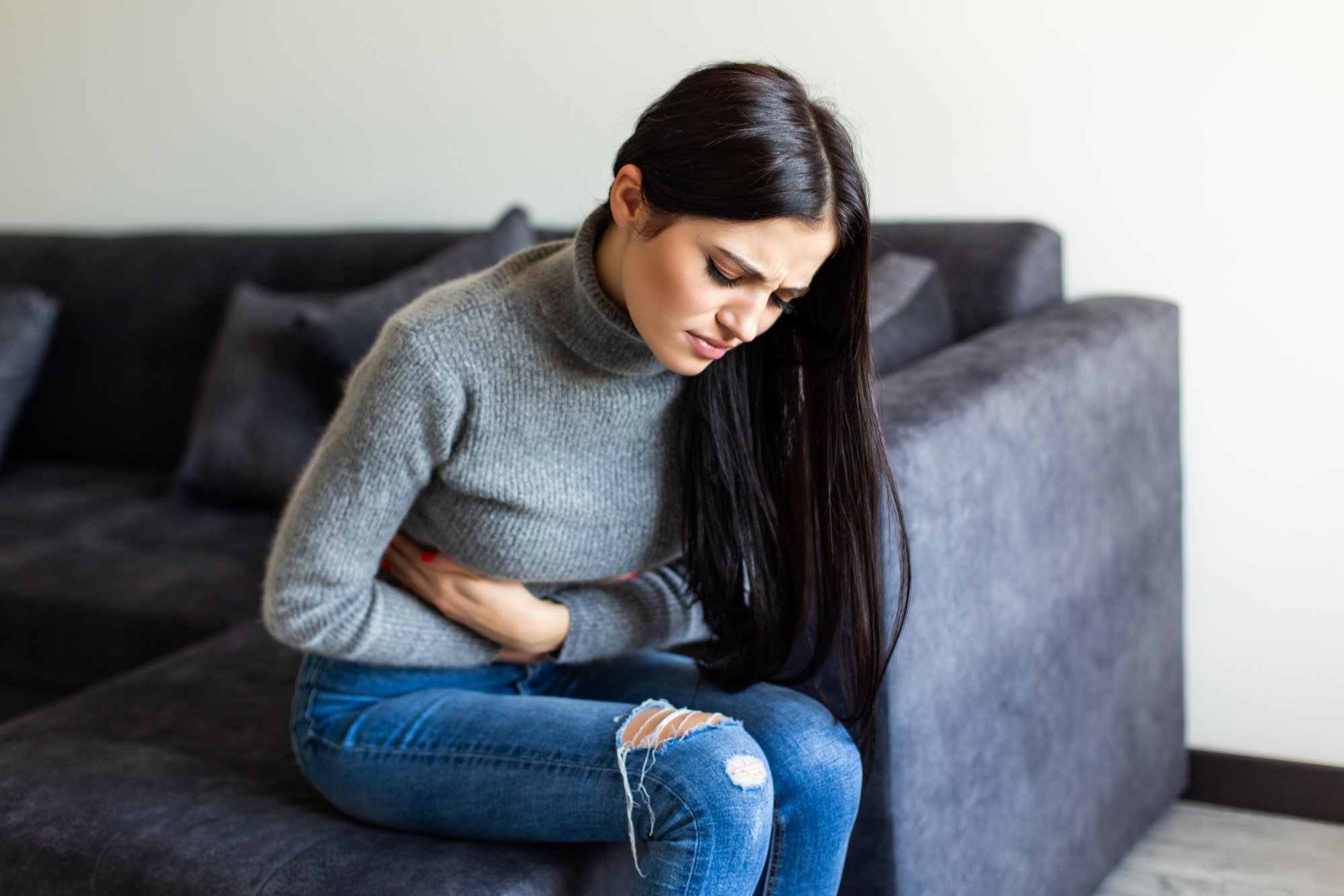 Eine Frau mit langen, dunklen Haaren sitzt auf einem grauen Sofa und hält sich den Bauch, offensichtlich unwohl.