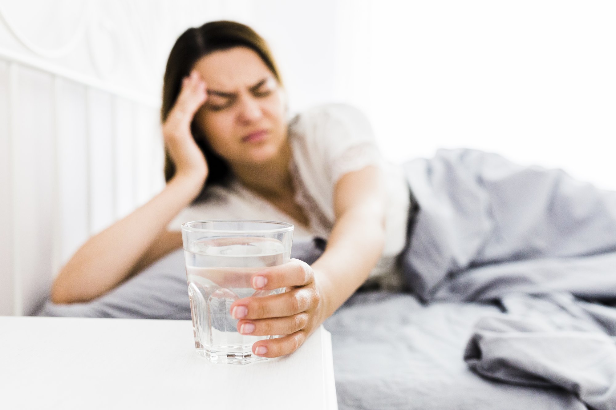 Eine Frau liegt im Bett und hält ein Glas Wasser, sichtbar unwohl und hat die Hand an die Stirn gelegt.
