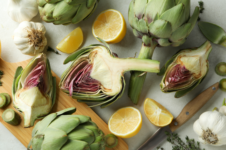 Frische Artischocken, einige halbiert, mit Zitronenscheiben und Knoblauch auf einem Brett angeordnet.