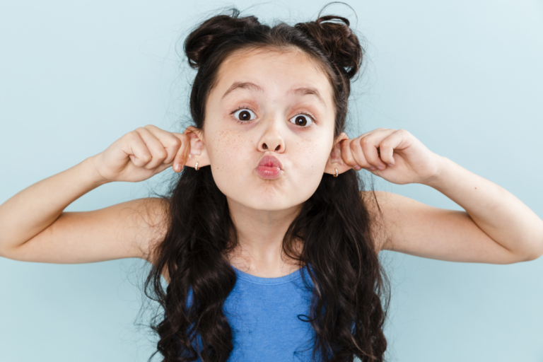 Ein Mädchen mit langen, lockigen Haaren zieht an ihren Ohren und macht ein lustiges Gesicht vor einer hellblauen Wand.