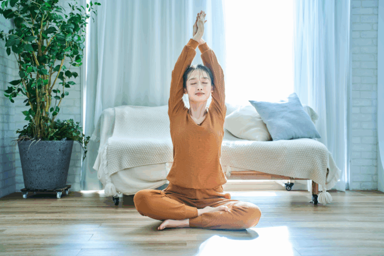 Eine Person in orangefarbenem Yoga-Outfit sitzt im Schneidersitz und streckt die Arme nach oben, umgeben von heller, luftiger Wohnungseinrichtung.