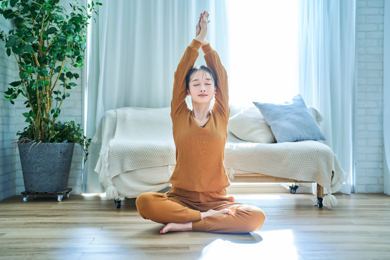 Vagusnerv.png Eine Person in orangefarbenem Yoga-Outfit sitzt im Schneidersitz und streckt die Arme nach oben, umgeben von heller, luftiger Wohnungseinrichtung.