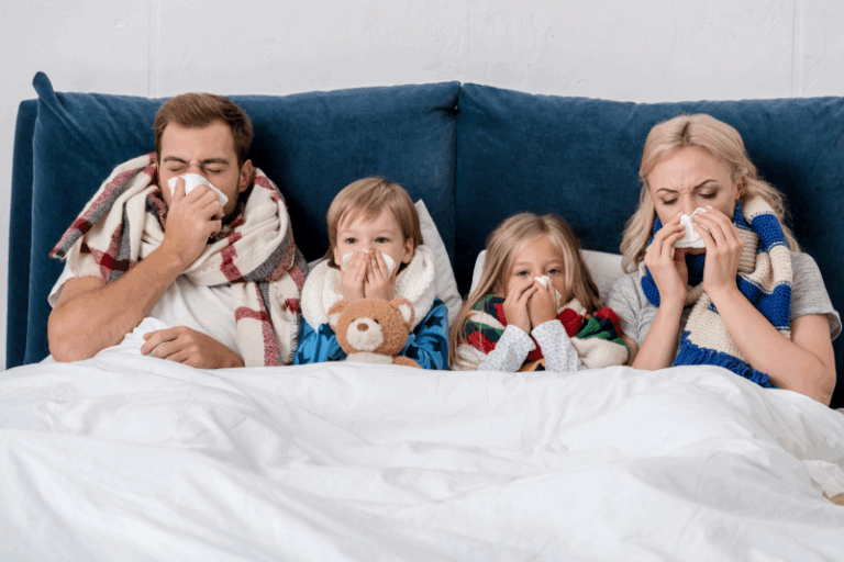 Eine Familie mit zwei Kindern kuschelt sich in Decken auf einer Couch, alle halten Taschentücher vor den Nasen.