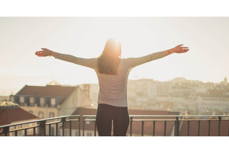 Eine Person mit ausgestreckten Armen steht auf einem Balkon und blickt in die Sonne über einer Stadt.