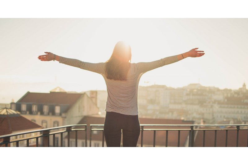 Eine Person mit ausgestreckten Armen steht auf einem Balkon und blickt in die Sonne über einer Stadt.
